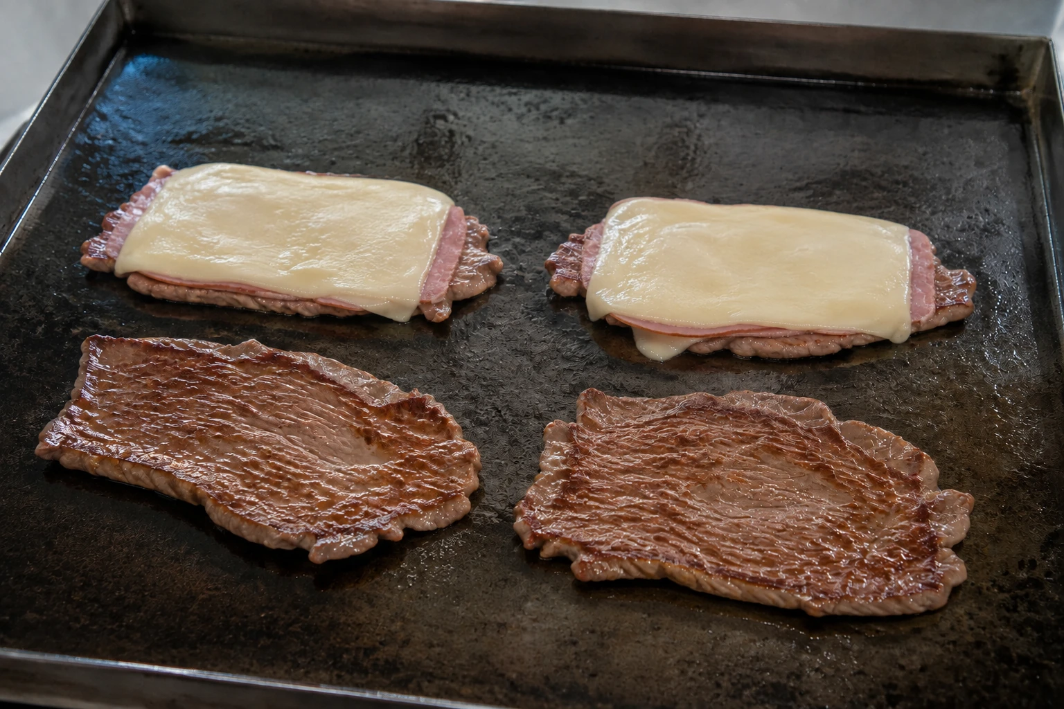 Four thin beef cutlets on a flat griddle, two topped with cooked ham and melting pale cheese