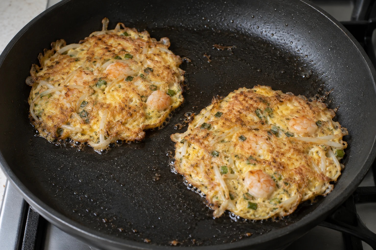 Two separate golden shrimp and bean sprout omelettes shallow frying in a black skillet