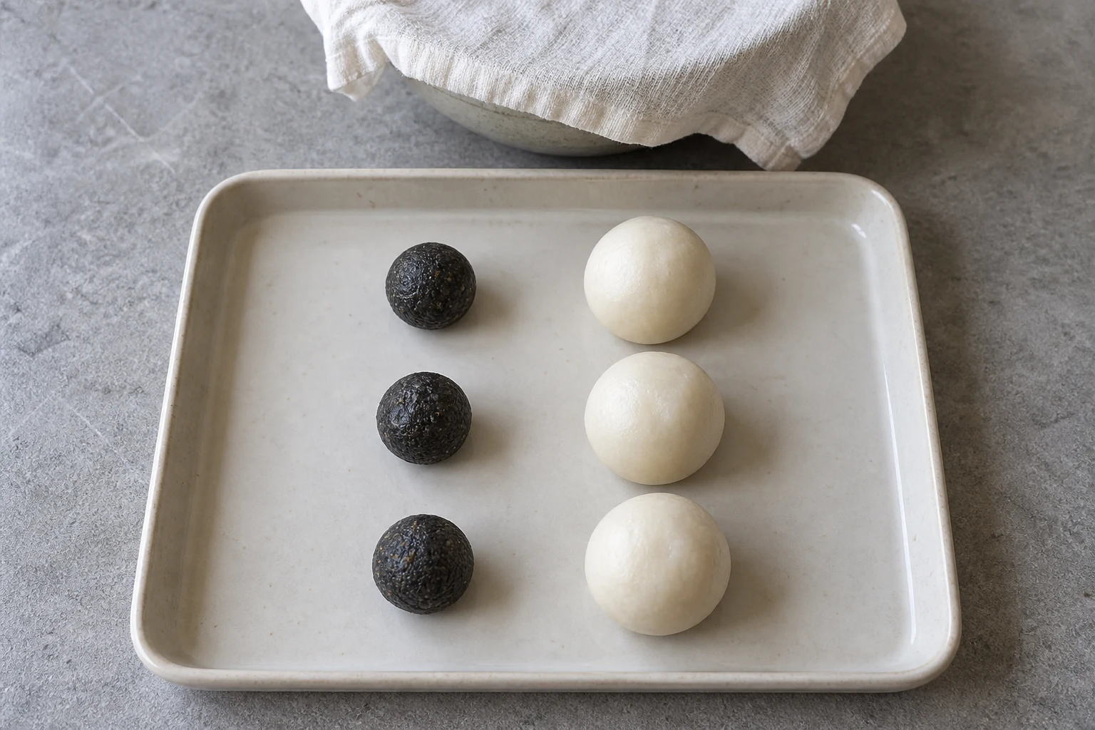 Three small dark sesame filling portions beside three larger white rice dough portions, with a covered bowl behind