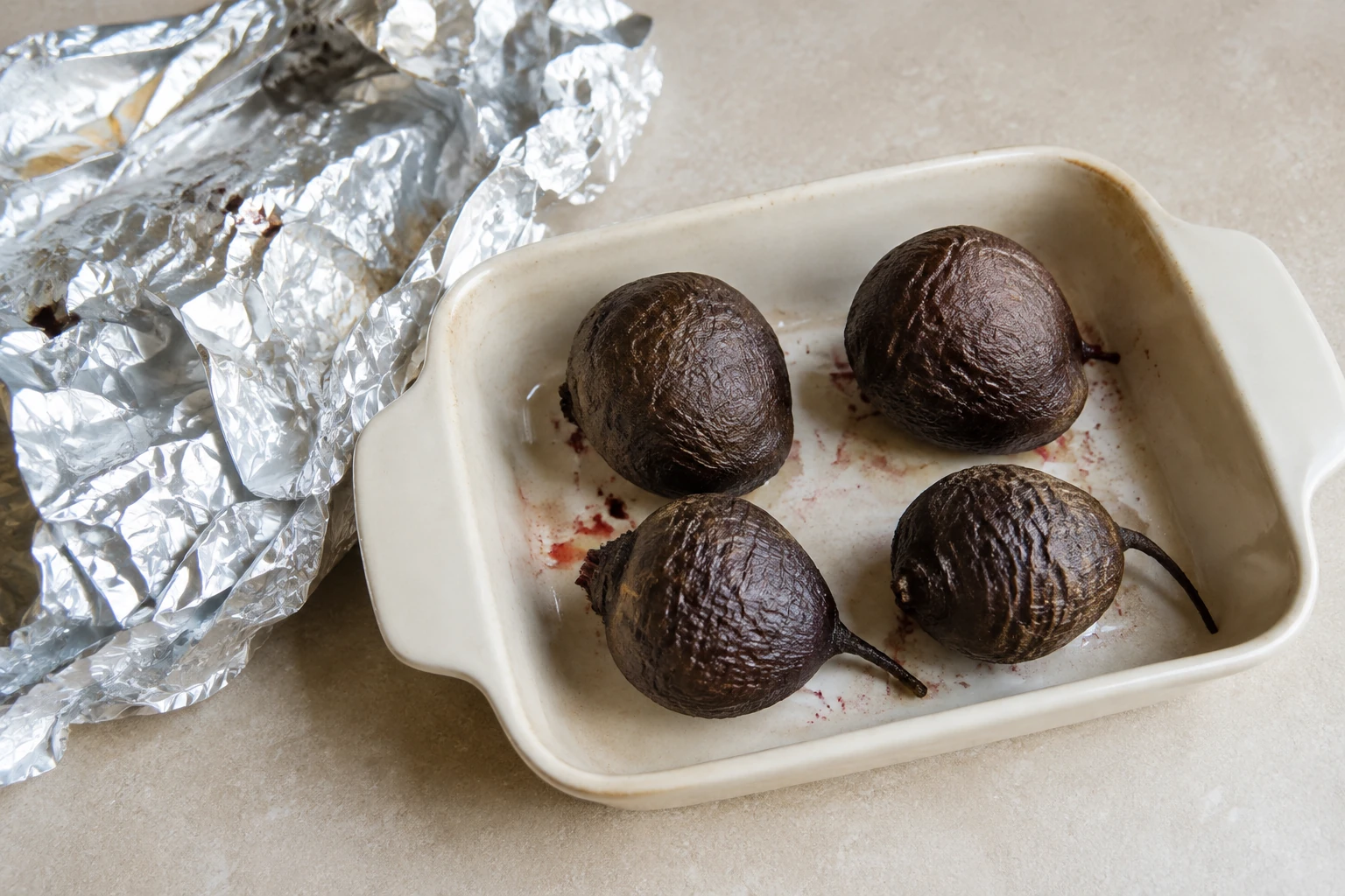 Four whole roasted beets with wrinkled skins in a ceramic baking dish beside opened foil