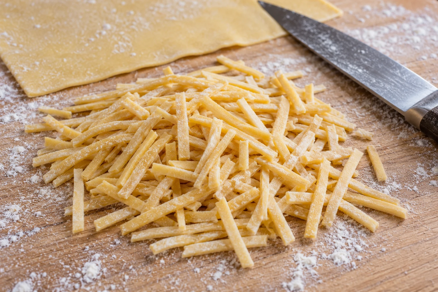 Short solid hand-cut yellow egg-noodle strips on a floured board beside rolled dough and a knife.