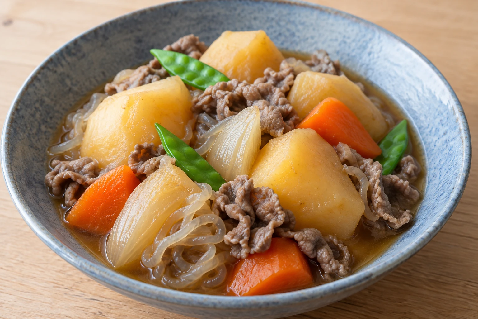 A bowl of nikujaga with golden potato chunks, thin beef slices, carrot, onion, shirataki and snow peas