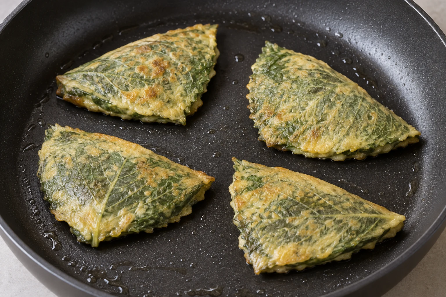 Four folded perilla-leaf packets spaced apart in a lightly oiled frying pan