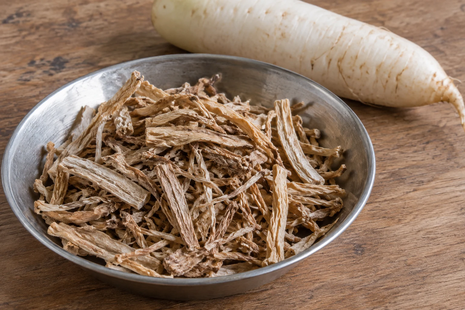 A bowl of wrinkled tan dried radish slivers with a fresh white radish root behind