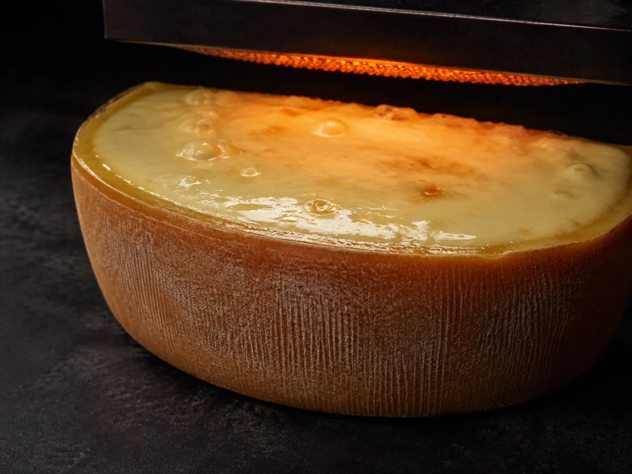 The pale cut face of a half wheel of raclette cheese softening beneath a heater